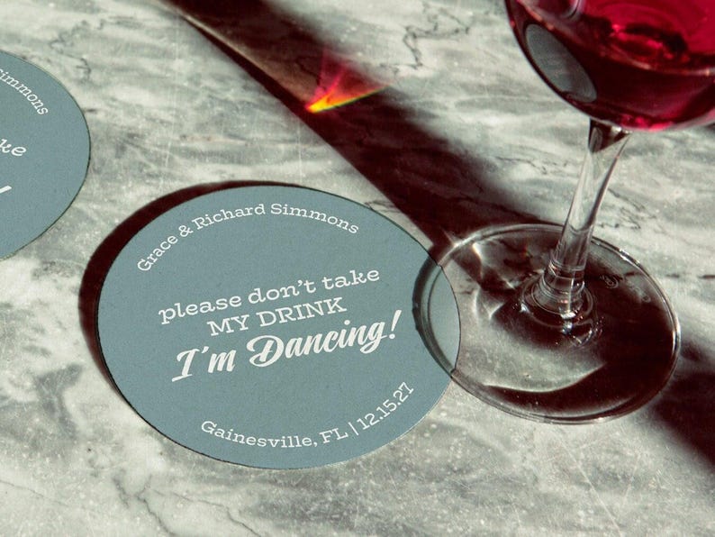 Coaster is shown with a wine glass on top of it and another off to the side. Coasters say Please dont take my drink, Im dancing with wedding couples names, location, and wedding date.