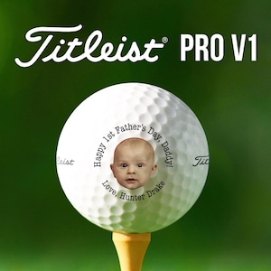 Top text reads Titleist Pro V1. Titleist golf ball is shown on a golf tee against a dark green background. Ball is shown with 1st Fathers Day photo design in black text color option.