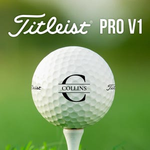 May include: A white Titleist PRO V1 golf ball with a dimpled surface, resting on a white tee. The ball features the black text COLLINS centered within a large letter C, and the Titleist brand name is printed on the sides of the golf ball.