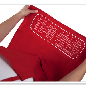 May include: Red kitchen towel with white text and a dotted border. The text includes baking measurements for different sizes of round and square pans, including 8-inch, 9-inch, 10-inch, and 12-inch.
