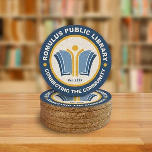 A stack of coasters sit on a wooden surface. The imagery on the coaster shows an open book with white writing, and blue and yellow art.