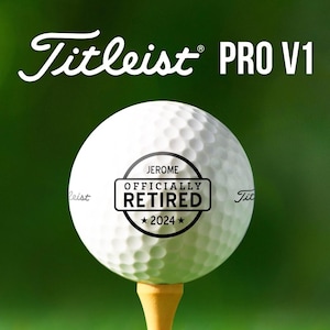 May include: A white Titleist PRO V1 golf ball with a black stamp that reads "JEROME OFFICIALLY RETIRED *2024*" on a green background.