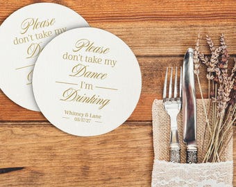 May include: Two round white coasters with gold text that reads: Please don't take my Dance I'm Drinking, followed by the names Whitney & Lane and the date 05/17/27. The coasters feature a smooth finish and are displayed alongside a silver fork and knife.