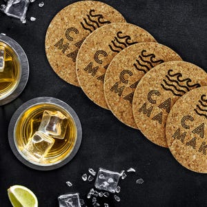 A set of four cork coasters sit on black surface with two drinks next to them. The coasters have Casa Makai text and wave imagery on it.