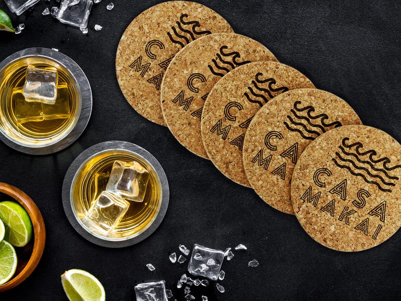 A set of four cork coasters sit on black surface with two drinks next to them. The coasters have Casa Makai text and wave imagery on it.