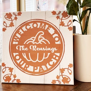 May include: A white canvas sign with a brown pumpkin-shaped circle that says "Welcome to The Rensings Our Patch". The sign has a white background with brown vines and leaves around the edges.