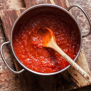 May include: A stainless steel pot filled with red tomato sauce, a wooden spoon with the text "Oldest Sunday Sauce Spoon" is in the pot.