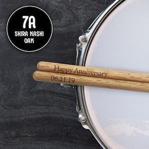 May include: A pair of wooden drumsticks with the text "Happy Anniversary 06.21.19" engraved on them. The drumsticks are resting on a silver drum.