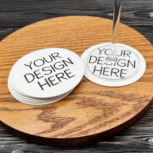 May include: A stack of circular white coasters and a single clear acrylic wine glass charm, both featuring the black printed text YOUR DESIGN HERE. The items are displayed on a round wooden surface with a visible grain pattern.