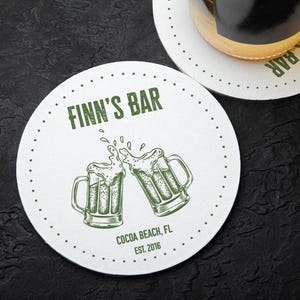 One coaster has a beer glass on it and an empty coaster sits beside it. Coasters feature bar design. This design uses green lettering on a white background, and a drawing of beer glasses. The name, established date, and location can be customized.