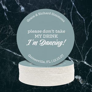 A single coasters sits on top of a stack of coasters against a dark marble background. Coaster says Please dont take my drink, Im dancing with wedding couples names, location, and wedding date.
