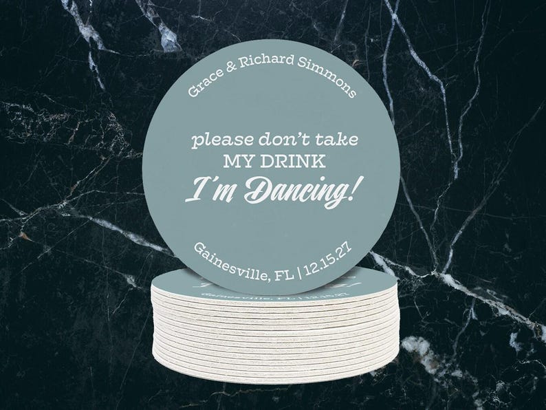 A single coasters sits on top of a stack of coasters against a dark marble background. Coaster says Please dont take my drink, Im dancing with wedding couples names, location, and wedding date.