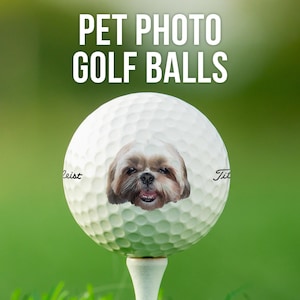 May include: A white golf ball with a dog's face printed on it. The dog is a Shih Tzu with brown fur and a white beard. The text "Titleist" is printed on the left side of the ball and "Title" is printed on the right side of the ball.