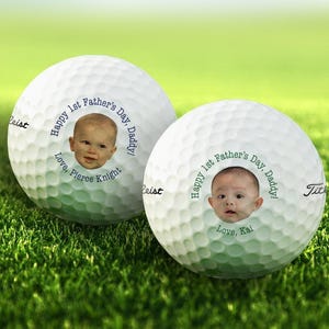 Titleist golf balls are shown on patch of grass. Both golf balls show 1st Fathers Day photo design in blue and green text color options.