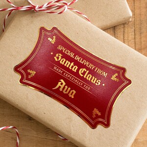 A gold foil cardstock Santa gift tag is shown on a craft paper wrapped box. The box also has a red and white striped string on it. The box is seen on a wooden surface.