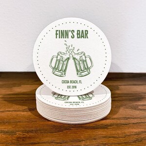 Single coaster sits on top of a stack of coasters. Coasters feature bar design. This design uses green lettering on a white background, and a drawing of beer glasses. The name, established date, and location can be customized.