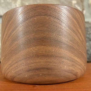 May include: A handcrafted, cylindrical wooden bowl with a smooth, curved design. The bowl is made of dark brown wood, showcasing the natural grain patterns. The bowl sits on a wooden surface.