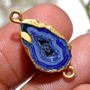 May include: A blue and gold gemstone pendant. The pendant features a polished, blue agate stone with a gold-coloured edge and two gold loops for attaching to a chain or other jewellery. The stone has a geode-like appearance with concentric blue bands.