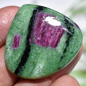 May include: A polished gemstone with a green base, featuring vibrant magenta and black inclusions. The stone has a smooth, rounded shape, with a prominent magenta area in the center and smaller magenta spots throughout. The black inclusions create a striking contrast.