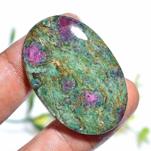 Puede incluir: Gema ovalada con base verde e inclusiones de rubí rosa. La superficie pulida revela un patrón natural y texturizado. Los cristales de rubí están dispersos por toda la piedra.