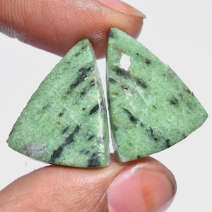 以下が含まれることがあります： 指で挟まれた、黒い縞模様のある緑色の三角形の宝石が2つ。石はざらざらとした質感の表面と光沢のある外観をしています。宝石は、ジュエリー作りやコレクションに使用される可能性があります。
