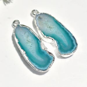 May include: Two agate pendants with a silver-coloured metal trim. The pendants have a teardrop shape with a light blue and teal colour scheme. The pendants have a loop at the top for hanging.
