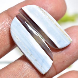 May include: Two polished, elongated gemstones with a light blue and brown striped pattern. The stones have a rounded shape and are held in a hand, showcasing their natural beauty and unique colour variations. The gemstones are likely used for jewellery or decorative purposes.