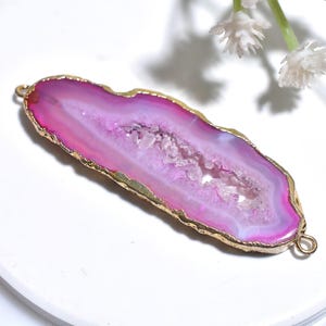 Puede incluir: Un colgante de ágata rosa y blanca con un borde dorado. La piedra tiene un centro similar a una geoda y dos pequeños bucles para sujetarlo a una cadena u otro componente de joyería. El colgante está sobre un fondo blanco.