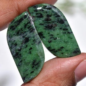 May include: Two polished, teardrop-shaped gemstones. The stones are a vibrant green with black speckles and streaks. The gemstones are held between fingers, showcasing their smooth, reflective surfaces. The overall aesthetic is natural and organic.