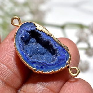 May include: A blue geode pendant with a gold-coloured trim and two loops for attaching to a chain. The geode has a deep blue interior with a textured surface, and the gold trim has a natural, irregular edge. The pendant is held in a hand.