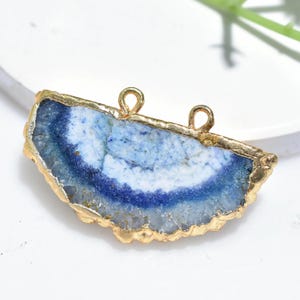 Puede incluir: Un colgante de ágata azul y blanca con un borde irregular dorado. El colgante tiene dos pequeños bucles en la parte superior para sujetarlo a una cadena o cordón. La piedra tiene un aspecto en capas con tonos de azul y blanco.