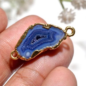May include: A blue and white agate gemstone pendant with a gold-coloured metal edge and two loops. The stone has a layered pattern and a central cavity. The pendant is held in a person's hand.