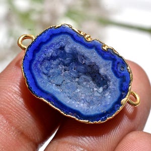 May include: A blue geode pendant with a gold-coloured trim and two small loops. The geode has a dark blue outer edge, a lighter blue interior, and a crystalline centre. The pendant is held by a person's fingers.