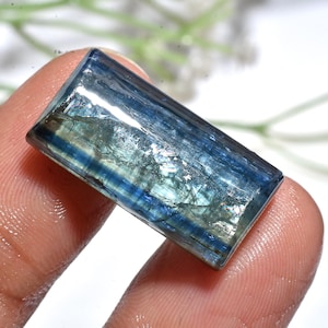 May include: A rectangular, polished gemstone displaying a gradient of blue shades. The stone is translucent, with visible internal structures and a reflective surface. The gemstone is held between fingers, highlighting its size and shape.