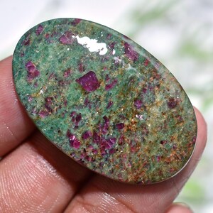 May include: Oval-shaped gemstone with a green base color, speckled with vibrant red ruby crystals. The stone has a polished surface, showcasing the natural patterns and colors of the minerals. The gemstone is held in a hand.