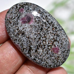 May include: An oval gemstone with a speckled grey and black surface, featuring several vibrant red ruby inclusions. The polished stone is held in a hand, showcasing its natural beauty and unique pattern.