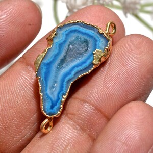 May include: A blue and white agate pendant with a gold-coloured trim. The pendant has an irregular shape and two small loops for attaching to a chain or other jewellery component. The agate's interior features a crystalline structure.