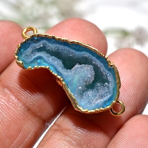 May include: A blue geode pendant with a gold-coloured trim and two small loops. The geode has a crystalline interior and a gold-coloured edge. The pendant is held in a hand.