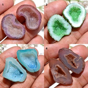 May include: Four pairs of polished geode halves in various colors. The geodes are in shades of purple, green, blue, and brown, with crystal formations visible inside. Each geode is held in a hand, showcasing the natural beauty and unique patterns of the stones.