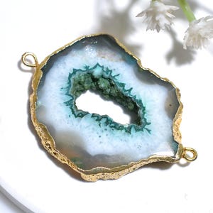 Pode incluir: Pingente de ágata natural com borda banhada a ouro. A pedra apresenta uma cavidade central com formações cristalinas verdes e brancas. Duas pequenas argolas douradas são fixadas para pendurar. Uma joia única.