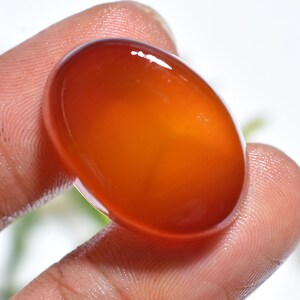 Puede incluir: Una gema pulida de forma ovalada, con tonos naranja y rojo. La piedra lisa tiene una superficie brillante y se sostiene entre dos dedos. La gema translúcida permite el paso de la luz, resaltando su color.