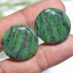 Könnte beinhalten: Zwei runde, polierte Zoisitsteine. Die Steine sind in einem lebendigen Grün mit schwarzen und violetten Adern gehalten. Die Steine werden in einer Hand gehalten, was ihre glatte, glänzende Oberfläche zeigt. Ideal für Schmuckherstellung oder Sammler.