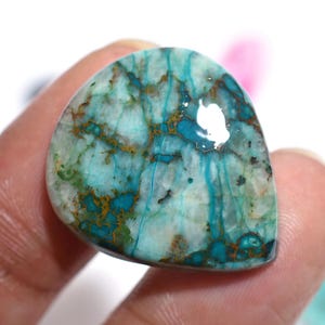 May include: A polished gemstone with a unique pattern of turquoise, teal, and brown veins. The stone has a smooth, rounded shape and a glossy finish. The colors blend together, creating a marbled effect. The gemstone is held in a person's hand.
