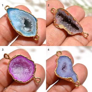 May include: Four gemstone pendants with gold-coloured edges and loops. The pendants feature different colours: light blue, lavender, magenta, and purple. The centre of each pendant has a crystal-like texture.