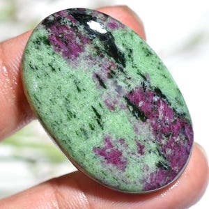 May include: Oval-shaped gemstone featuring a blend of colors. The stone showcases a light green base with streaks of deep magenta and black. The polished surface reflects light, highlighting the natural patterns and textures of the mineral.