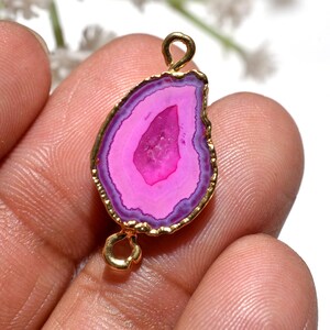 May include: A pink and purple agate gemstone pendant with a gold-coloured metal frame. The pendant has a teardrop shape with a gold loop at the top and bottom for attaching to a chain or other jewellery components.