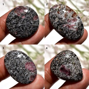 Peut inclure: Quatre pierres précieuses polies avec un motif moucheté gris et noir, chacune présentant une inclusion rouge foncé proéminente. Les pierres varient en forme, y compris des formes ovales et en goutte, et sont tenues dans une main.