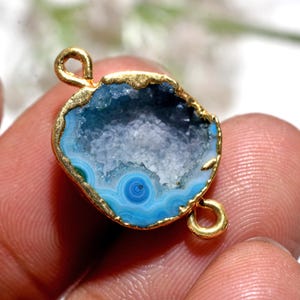 May include: A gold-plated pendant with a blue geode and a crystalline interior. The geode has a gold rim and two loops for attachment. The geode's interior features concentric blue patterns. The pendant is a jewellery component.
