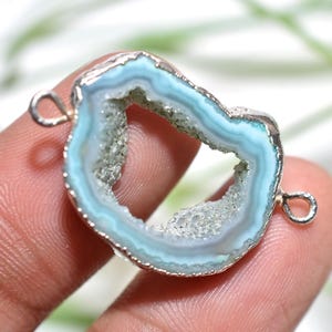 Op de afbeelding: Een lichtblauwe en zilveren hanger met een geode-ontwerp. De hanger heeft een zilverkleurig metalen frame en twee lussen om aan een ketting of andere sieraden te bevestigen. De geode heeft een lichtblauw en wit kleurenschema.