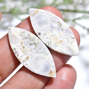 Puede incluir: Dos piedras preciosas pulidas, de forma ovalada, con aspecto veteado. Las piedras son principalmente blancas con inclusiones grises, beiges y verdes. Las piedras preciosas tienen un acabado liso y brillante.